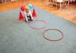 Dziewczynka w różwoej bluzce pokonuje tor przeszkód na niebieskim dywanie, z zaangażowaniem i radością uczestniczy w aktywności ruchowej