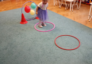 Dziewczyna w fioletowej sukience skacze po okrągłych przeszkodach na niebieskim dywanie, doskonaląc swoją równowagę i koordynację ruchową.