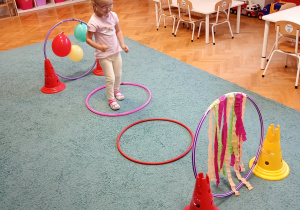 Dziewczyna w różowej bluzce skacze po okrągłych przeszkodach na niebieskim dywanie, doskonaląc swoją równowagę i koordynację ruchową.