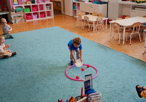 Chłopiec w niebieskiej bluzce niesie na talerzyku piłkę do okragłej obręczy na niebsiekim dywani