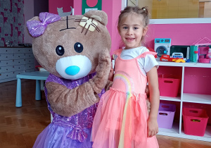 Milana z uśmiechem pozuje do zdjęcia z misiem ubranym w fioletową spódniczkę i kokardkę