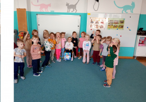 Wszytskie dzieici stoją na niebieskim dywanie. Na środku siedzi nasza solenizantka z kolorowa torebką ze słodkościami.