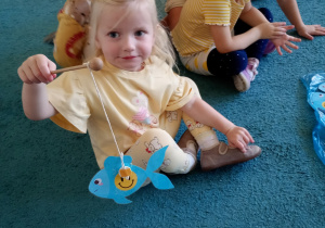 Dziewczyna w żółtej bluzce siedzi na niebieskim dywanie. Trzyma wedkę z rybką, na której jest uśmiech