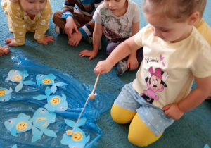 Dziewczyna siedzi na niebskiem dywanie. Łowi rybki z uśmiechem rozłożne na dywanie.