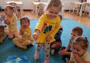 Uśmeichnięta dziewczyna w żółtej bluzce stara się złowić rybki rozłożone na niebieskim dywanie. W ręku trzyma wękę.