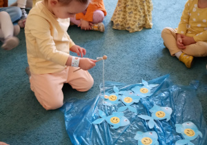 Dziewczynka w żółtej bluzce ze skupieniem łowi rybki rozłożone na niebsiekim dywanie.