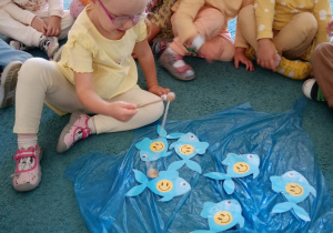 Dziewczyna w okularach ze skupieniem łowi rybki rozłożone na niebskiem dywanie.