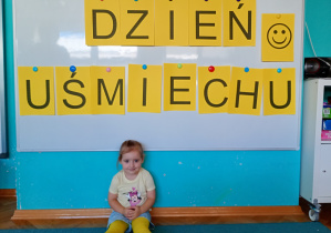 Dziewczyna w żółtych rajstopach siedzi na dywanie. Na tle napisu dzień uśmeichu przywieszonego na białej tablicy