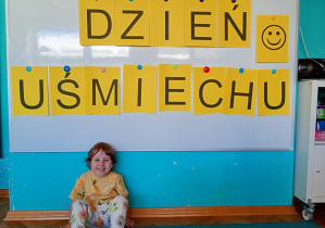 Uśmiechnięty chłopiec siedzi na niebieskim dywanie. Na tle napisu dzień uśmiechu przywieszonego na białej tablicy