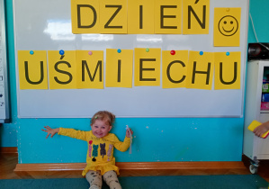 Dziewczynka w żółtej bluzce siedzi na niebskim dywanie. Na tle napisu dzień uśmiechu przywieszonego na białej tablicy