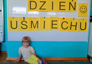 Dziewczynka w żółtych spodenkach siedzi na niebieskim dywanie. Na tle napisu dzień uśmiechu przywieszonego na białej tablicy