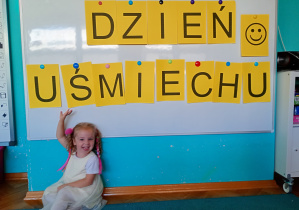 Dziewczynka w białej sukience siedzi na niebieksim dywanie. Na tle napisu dzień uśmiechu przywieszonego na białej tablicy.