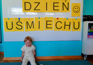 Dziewczynka siedzi na niebieskim dywanie. Na tle napisu dzień uśmiechu przywieszonego na białej tablicy