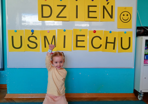 Dziewczynka w żółtej bluzce, z opaską na ręku siedzi na niebskim dywanie. Na tle napisu dzień uśmiechu przywieszonego na białej tablicy