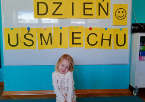 Dziewczynka siedzi na niebsiekim dywanie. Na tle napisu dzień uśmiechu przywieszonego na białej tablicy
