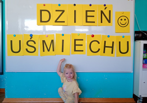 Dziewczyna w żółtej bluzce w jasnych włsach siedzi na niebskim dywanie. Na tle napisu dzień uśmiechu przywieszonego na białej tablicy