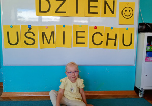 Dziewczynka w żółtej bluzce. Na tle napisu dzień uśmiechu przywieszonego na białej tablicy