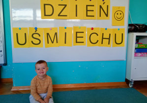 Chłopiec siedzi na niebskime dywanie. Na tle napisu dzień uśmiechu przywieszonego na białej tablicy