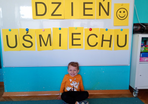 Chłopiec w pomarańczowej bluzce siedzi na niebieskim dywanie. Na tle napisu dzień uśmiechu przywieszonego na białej tablicy