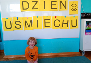 Dziewczynka w pomarańczowej bluzce siedzi na niebskiem dywanie. Na tle napisu dzień uśmiechu przywieszonego na białej tablicy