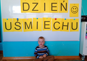 Chłopiec w bluzce w paski siedzi na niebskim dywanie. Na tle napisu dzień uśmiechu przywieszonego na białej tablicy