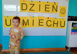 Chłopiec stoi na niebksim dywanie. Na tle napisu dzień uśmiechu przywieszonego na białej tablicy