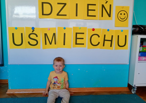 Chłopeic w żółtej koszulce siedzi na niebskiem dywanie. Na tle napisu dzień uśmiechu przywieszonego na białej tablicy