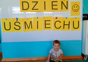 Chłopiec w jasnej koszulce siedzi na niebskim dywanie. Na tle napisu dzień uśmiechu przywieszonego na białej tablicy