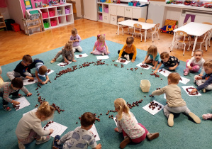 Dzieci siedzą w kole na niebieskim dywanie. Przed sobą mają białe kartki, w koło rozłożone sa kasztany.