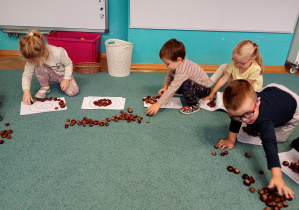 Dzieci siedza na niebskiem dywanie. Układają wzór z kasztanów na białych kartkach
