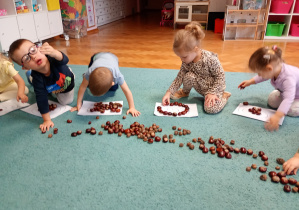 Dzieci siedza na niebskiem dywanie. Układają wzór z kasztanów na białych kartkach