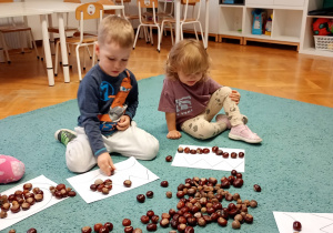 Dzieci siedza na niebskiem dywanie. Układają wzór z kasztanów na białych kartkach. Przed dzieićmi na dywanie leżą kasztany