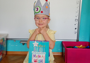 Solenizantka Jagoda w koronie na głwoie z cyfrą 3. W ręku trzyma niebieską torbę z tortem.