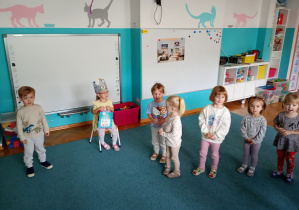 Dzieci stoją na niebieskim dywanie. Na środku siedzi solenizantka, trzyma w ręku niebieską torbę