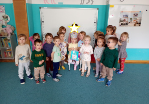 Wspólne zdjęcie dzieici, z solenizantką. Dzieci stoją na niebieskim dywanie. Na środku siedzi solenizantka w koronie na głowie.