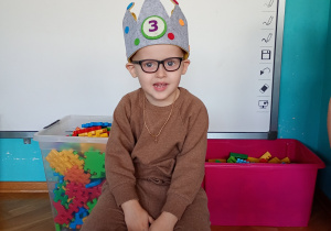 Nasz solenizant w koronie na głwoie siedzi na krześle. Przed sobą trzyma zieloną torbę z upiminkami dla dzieici.