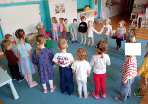 Dzieci stoją w kole na niebieskim dywanie. Solenizant siedzy na środku koła w koronie na głowie.