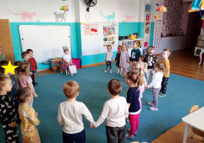 Dzieci stoją wkole na niebsiekim dywanie. Na środku, na krześle siedzi nasza solenizantka.