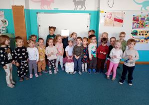 Dzieci stoją na niebsiekim dywanie. Na środku siedzi solenizantka, w rękach trzyma torebkę w kolorowe serduszka.