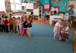 Dzieci stoją w kolejce na niebsiekim dywanie. Na krześle przed dzieźmi siedzi nasza solenizantka. Dzieci podchodza po kolei, składają życzenia i czestują się słodościami.