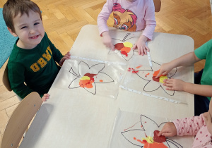 Ignacy i Ala siedzą przy stoliku. Przed soba mają folię z farbą w środku. Ala rozporowadza farbę palcem po konturze.