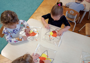 Dzieci siedzą przy stoliku. Na stolikach lezą folie z farbami i konturami liścia. dzieci rozporwadzaja paluszkiem farby, maluja liście.