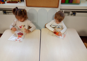 Dzieci siedzą przy stoliku. Na stolikach lezą folie z farbami i konturami liścia. dzieci rozporwadzaja paluszkiem farby, maluja liście.
