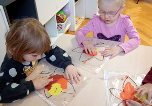 Dzieci siedzą przy stoliku. Na stolikach lezą folie z farbami i konturami liścia. dzieci rozporwadzaja paluszkiem farby, maluja liście.