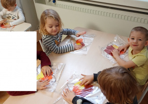 Dzieci siedzą przy stoliku. Na stolikach lezą folie z farbami i konturami liścia. dzieci rozporwadzaja paluszkiem farby, maluja liście.