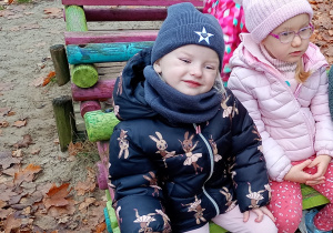 Dzieci w kolorywhc kurtkach, czapkach bawią się w pociągu. W tle broązowe, jesienne liście