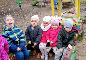 Dzieci w kolorywhc kurtkach, czapkach bawią się w pociągu. W tle biegają dzieci.