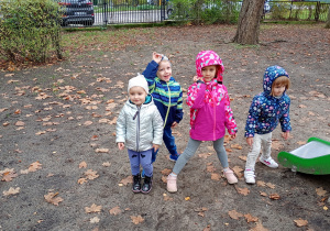 Dzieic w kolorywch kurtkach i czapkacch akwtynie sopędzają czas w przedszkolnym ogrodzie. W tle brązowe, jesienne liscie.