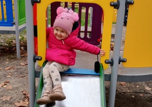 Matylda w różwoej czapce i kurtce, z uśmiechem na twarzy zjeżdża na zjeżdżalni.