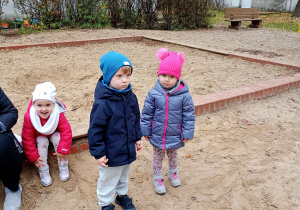 Przedszkolaki w kolorwych kurtkach i czapkach pozują do zdjęcia. W tle znajduje się piaskownica.